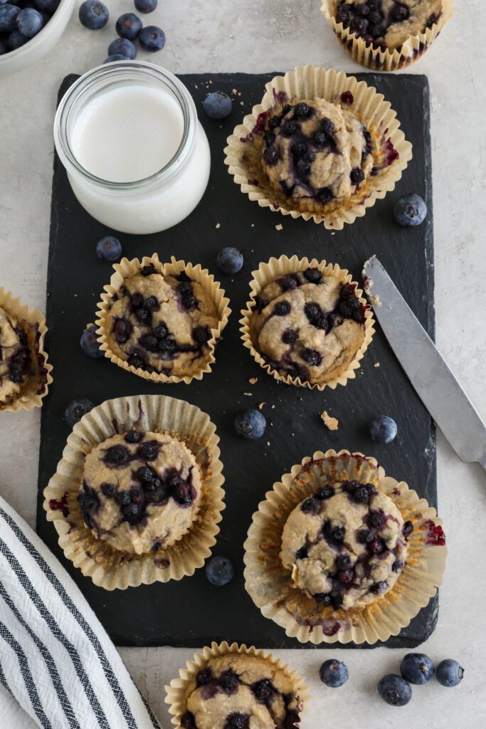 Vegan Blueberry Muffins served on black rectangle with glass of milk, knife, and blueberries 