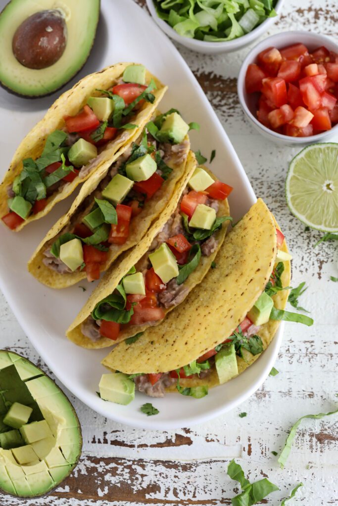 Vegan Refried Bean Tacos served on platter topped with diced tomato, lettuce, avocado, and cilantro 