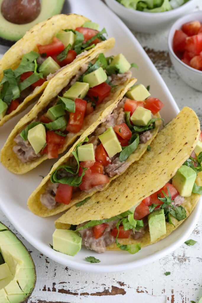 Vegan Refried Bean Tacos served on platter topped with diced tomato, lettuce, avocado, and cilantro 