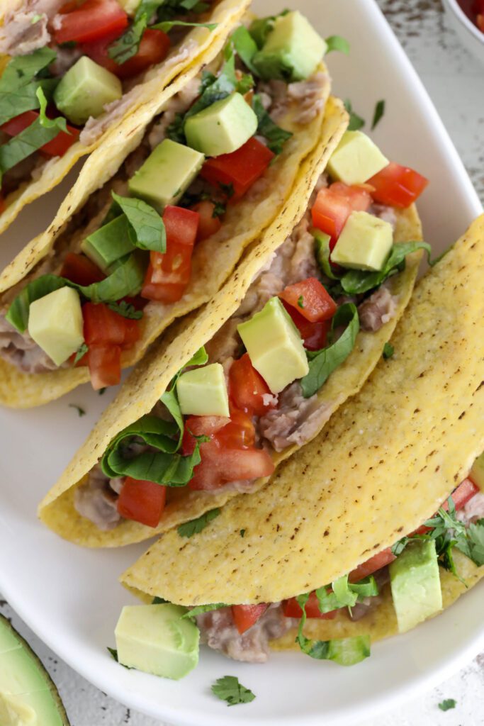 Vegan Refried Bean Tacos served on platter topped with diced tomato, lettuce, avocado, and cilantro 
