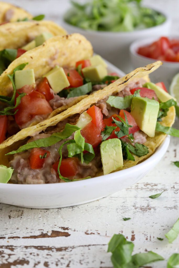 Vegan Refried Bean Tacos served on platter topped with diced tomato, lettuce, avocado, and cilantro 