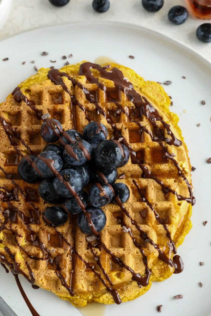 Pumpkin Turmeric Waffles served on plate topped with blueberries, cacao nibs, and chocolate drizzle 