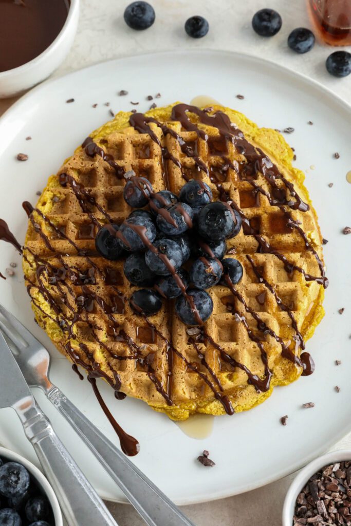 Pumpkin Turmeric Waffles served on plate topped with blueberries, cacao nibs, and chocolate drizzle 