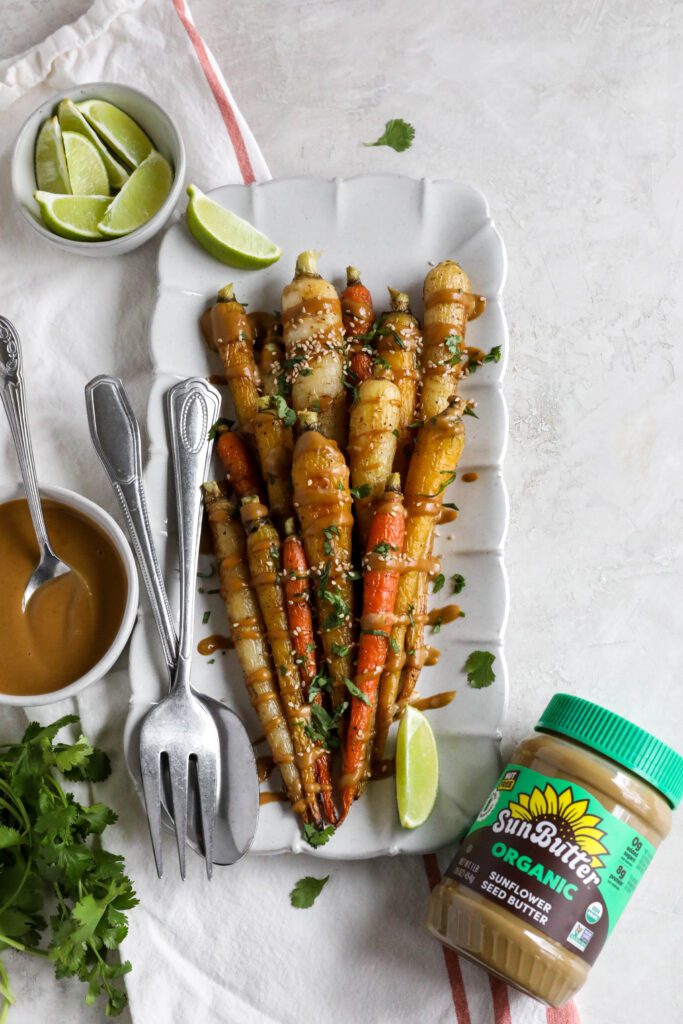 Rainbow Roasted Carrots with SunButter Sauce served on rectangular platter with fork and spoon topped with lime and cilantro and sesame seeds