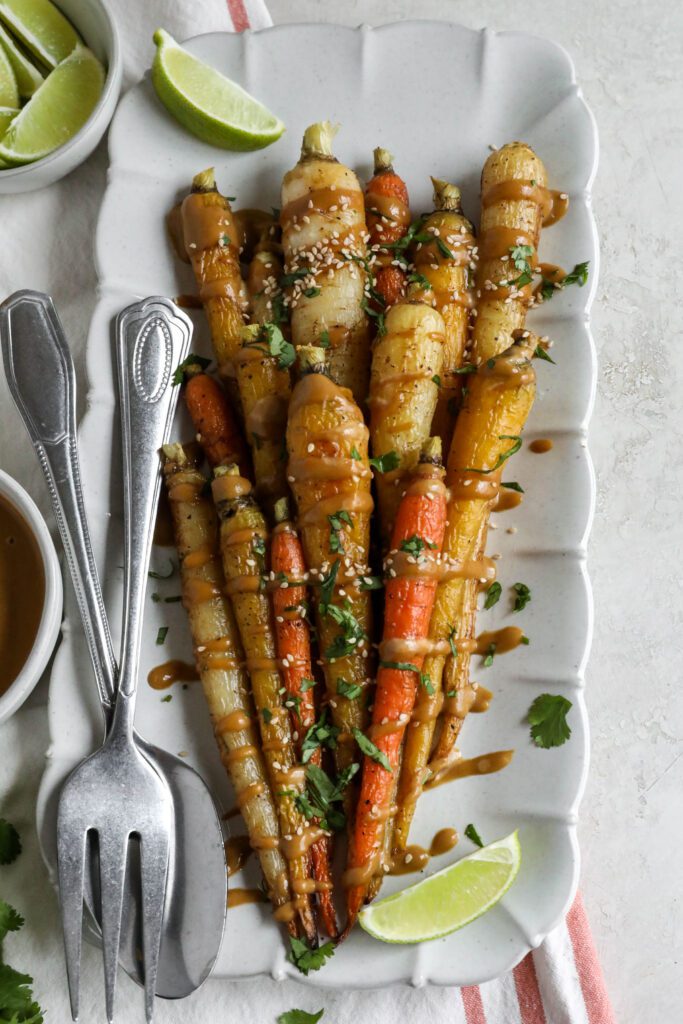 Rainbow Roasted Carrots with SunButter Sauce served on rectangular platter with fork and spoon topped with lime and cilantro and sesame seeds