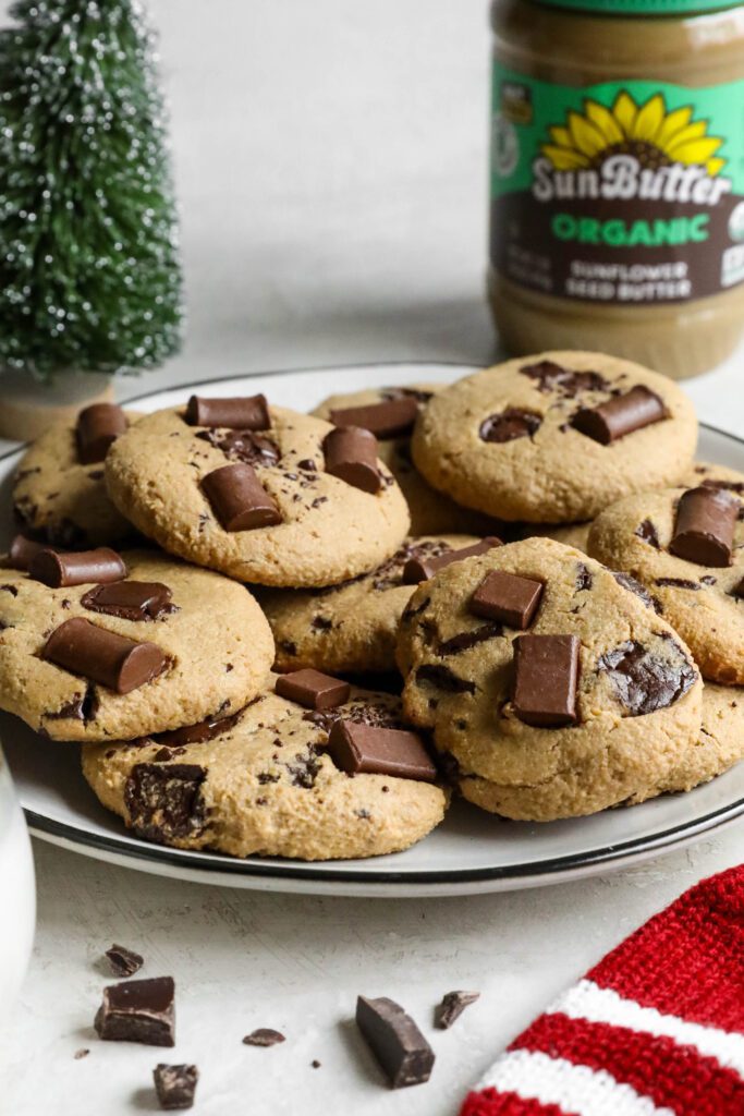 SunButter Chocolate Chip Cookies stacked on plate with glass of almond milk, santa hat, and evergreen tree