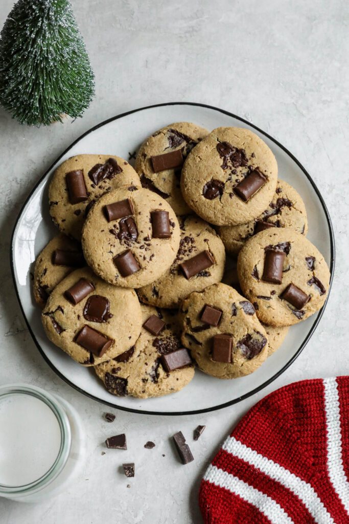 SunButter Chocolate Chip Cookies stacked on plate with glass of almond milk, stocking, and evergreen tree