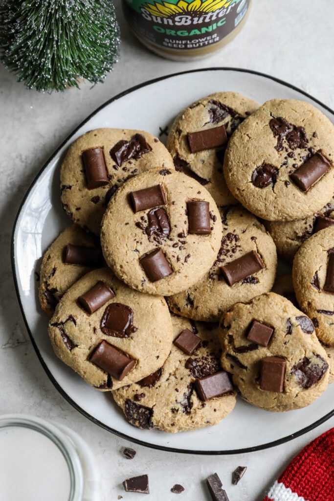 SunButter Chocolate Chip Cookies stacked on plate with glass of almond milk, santa hat, and evergreen tree