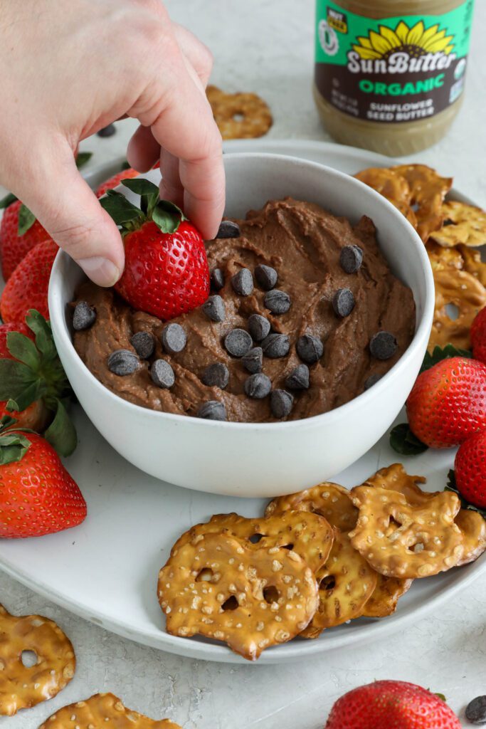 Chocolate SunButter Hummus served in bowl surrounded by plate of pretzels and strawberries with hand dipping strawberry