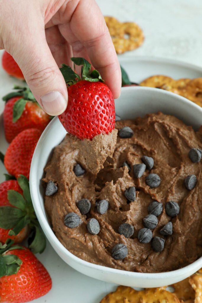 Chocolate SunButter Hummus served in bowl surrounded by plate of pretzels and strawberries with hand dipping strawberry