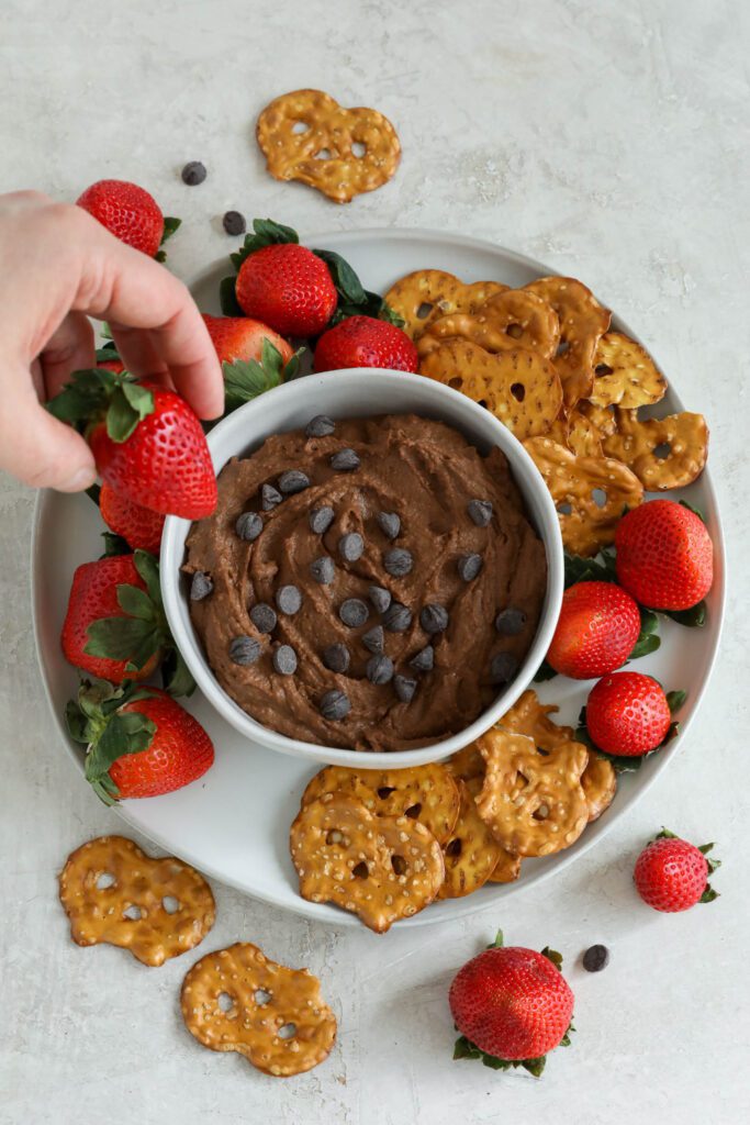 Chocolate SunButter Hummus served in bowl surrounded by plate of pretzels and strawberries with hand dipping strawberry