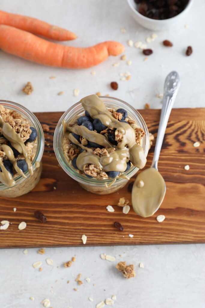 Carrot Cake SunButter Overnight Oats served in mason jars on wooden board topped with blueberries, granola, and SunButter 